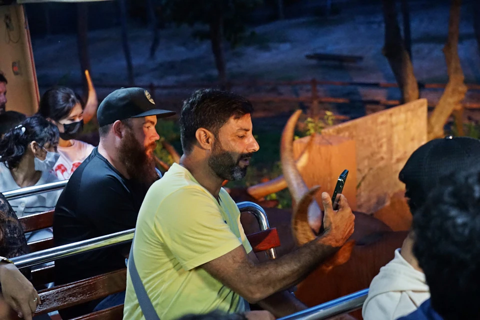 A man wearing a yellow shirt sits on a ride holding a phone, with another bearded man seated behind him in an outdoor setting.