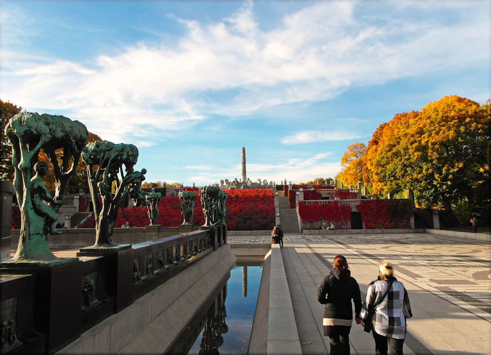 People walking along a pathway lined with sculptures and trees with autumn foliage, under a sunny blue sky.