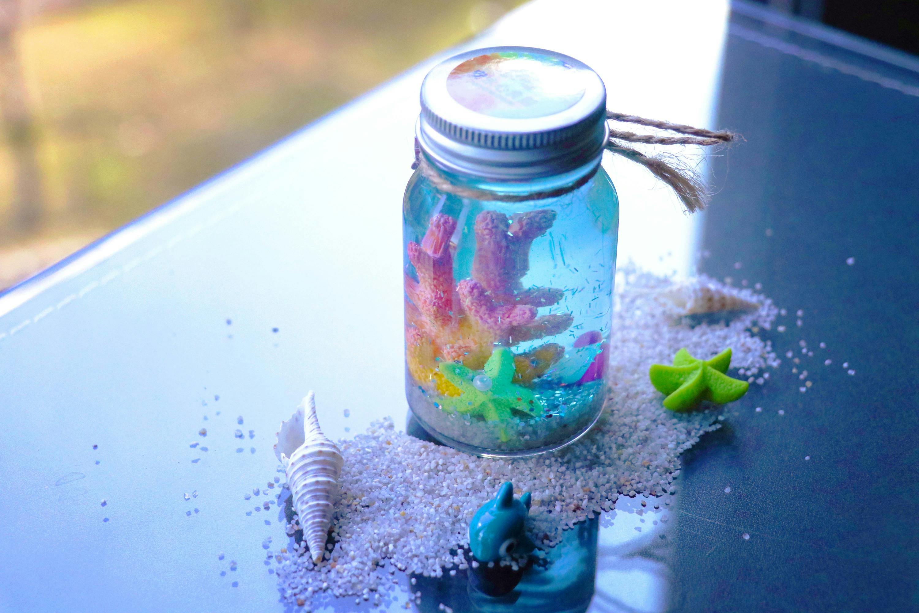 A glass jar with colorful coral-like decorations inside, placed on sand with small shells and a toy shark around it.