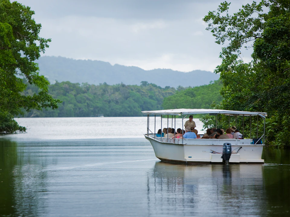 A boat with a canopy carries several people on a calm lake surrounded by lush greenery and misty mountains in the background.