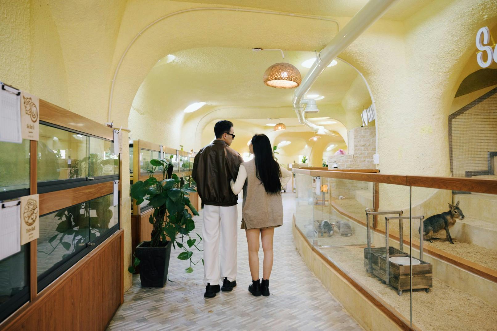 Two people walking together in a brightly lit, yellow-toned hallway with glass displays and potted plants.