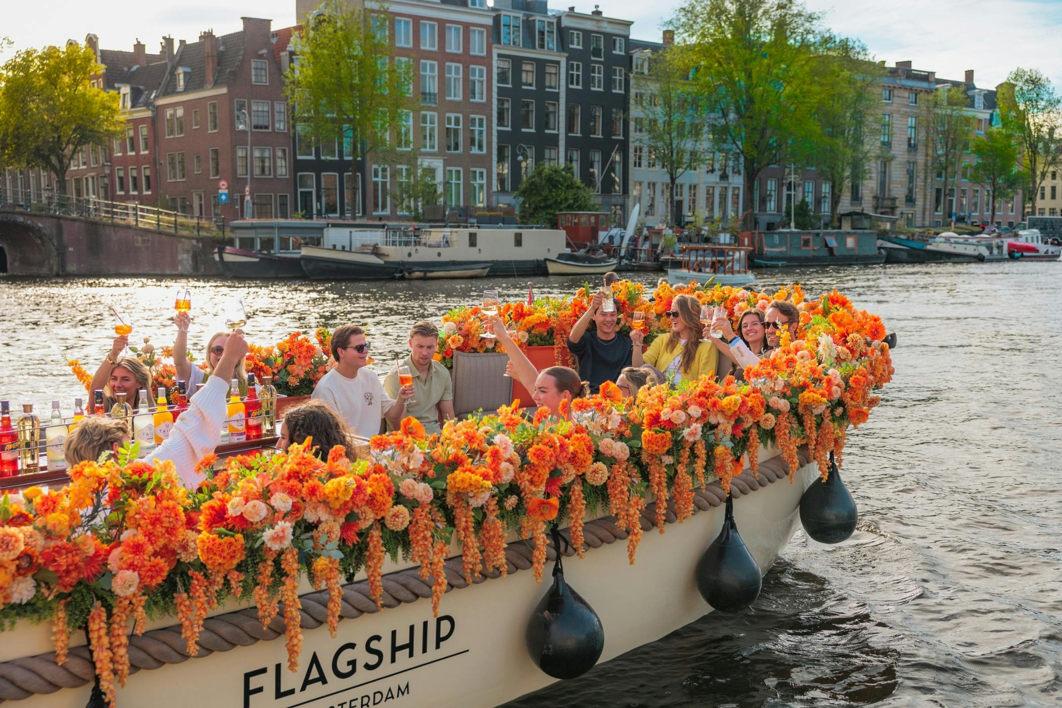A boat adorned with orange and yellow flowers carrying people enjoying drinks on a canal in a European city.