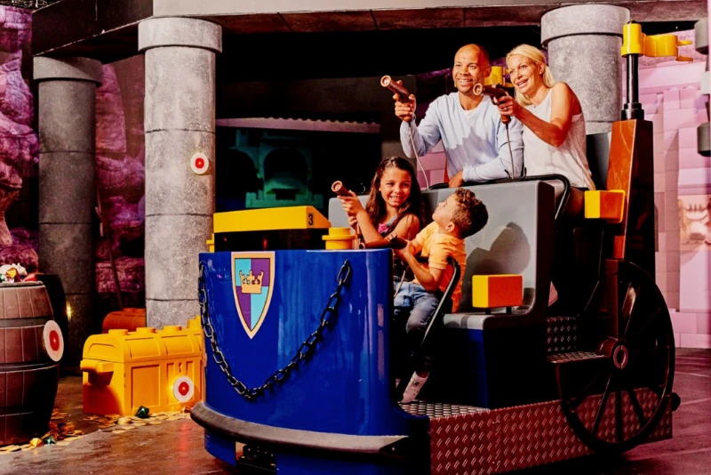 A family of four enjoys a ride, aiming toy guns on a blue cart with a shield emblem.