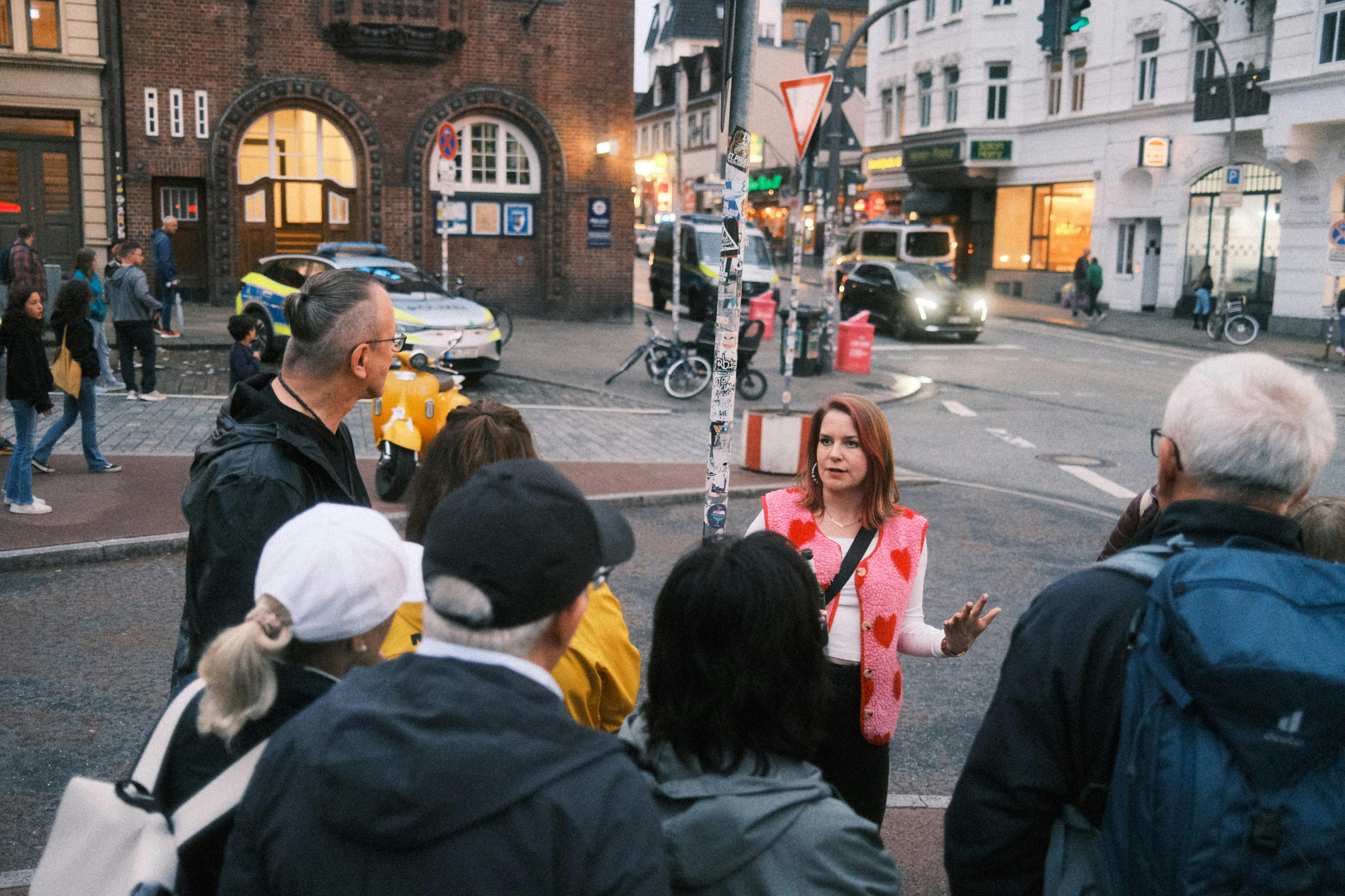 Μια γυναίκα με ροζ γιλέκο με σχέδιο καρδιάς οδηγεί μια ομάδα περιηγήσεων σε έναν πολυσύχναστο αστικό δρόμο με κτίρια, οχήματα και ποδήλατα στο παρασκήνιο.