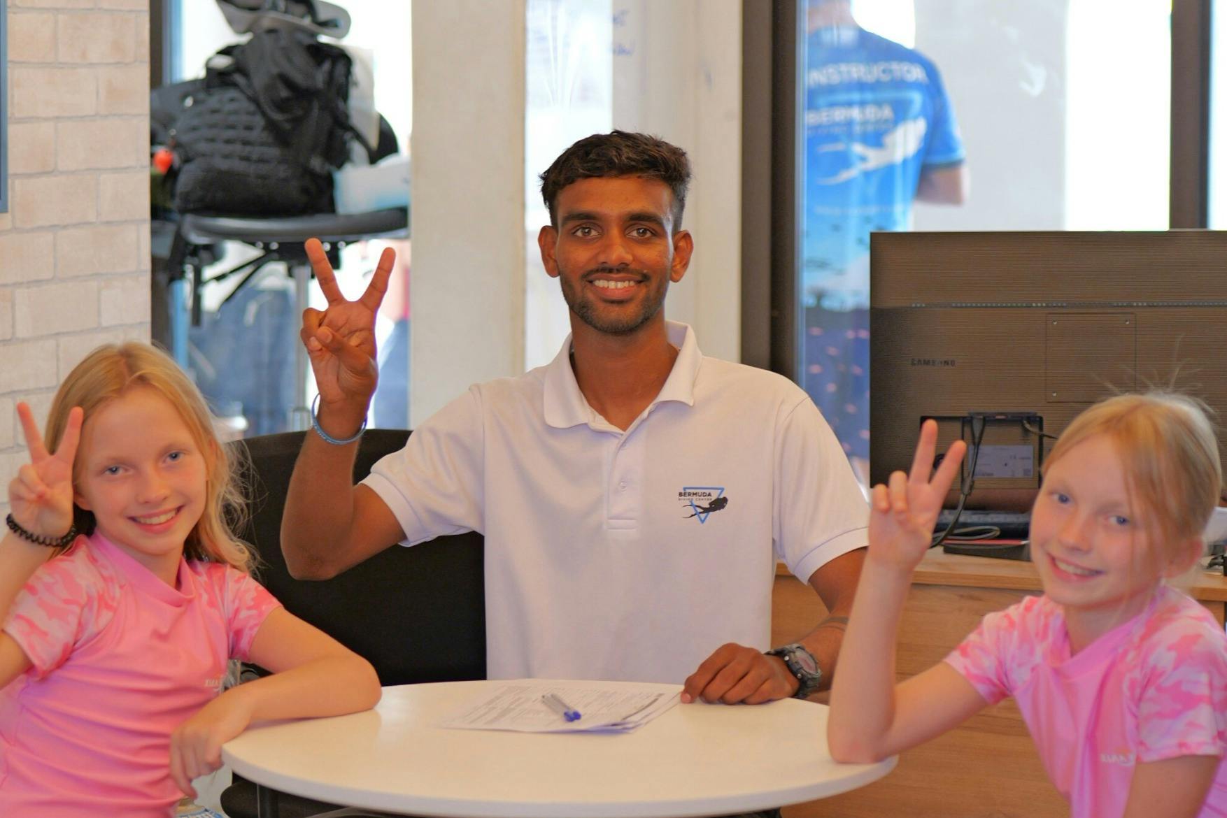 白いポロシャツを着た男性がテーブルに座り、ピースサインを掲げています。前景には2人の子供が写っています。