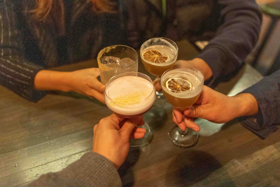 Four people clinking glasses with various drinks, garnished, over a wooden table in a social setting.