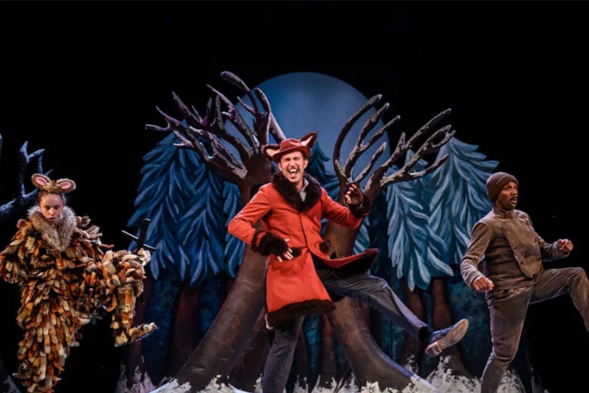 Tři herci v barevných kostýmech vystupují před lesní scénou s velkými malovanými stromy a měsíčním pozadím.