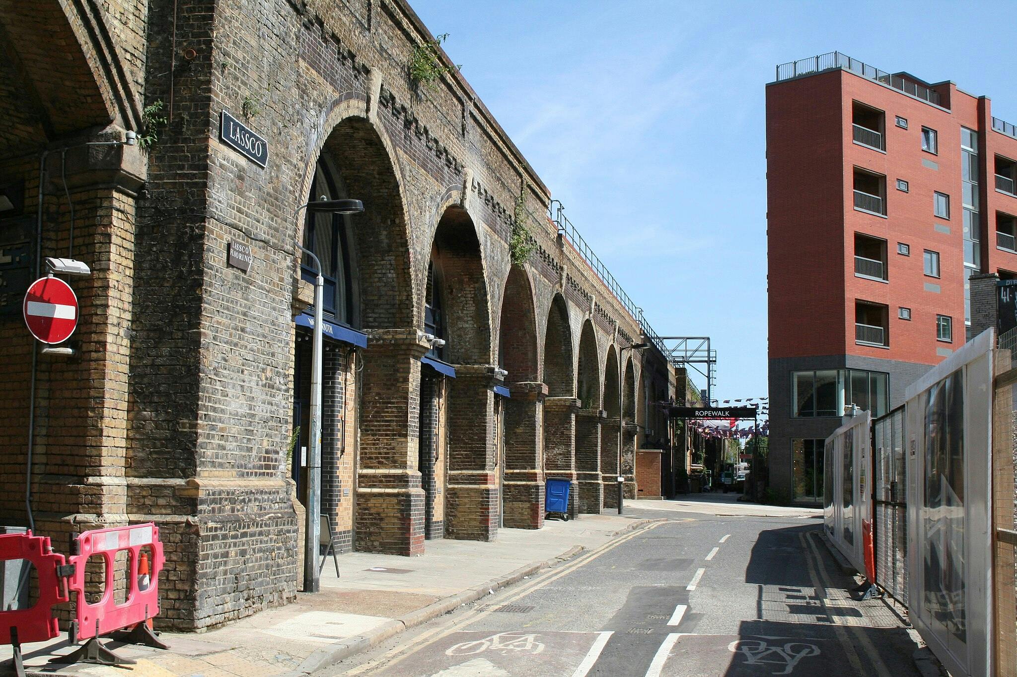 Arcades en pierre sur un viaduc en brique le long d'une rue où se trouve un bâtiment moderne de couleur rouge. Les panneaux indiquent "Lassco Ropewalk".