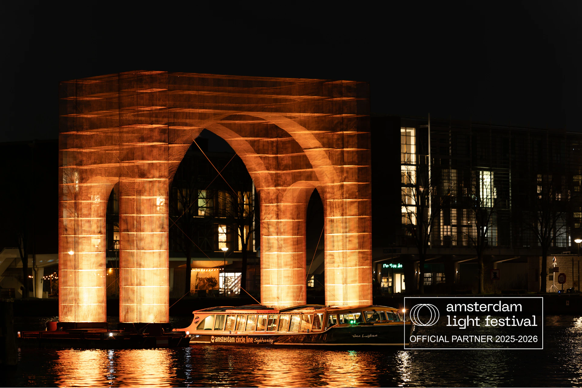 An illuminated arch sculpture over water at night, with a boat passing underneath and buildings in the background. Banner: "Amsterdam Light Festival".