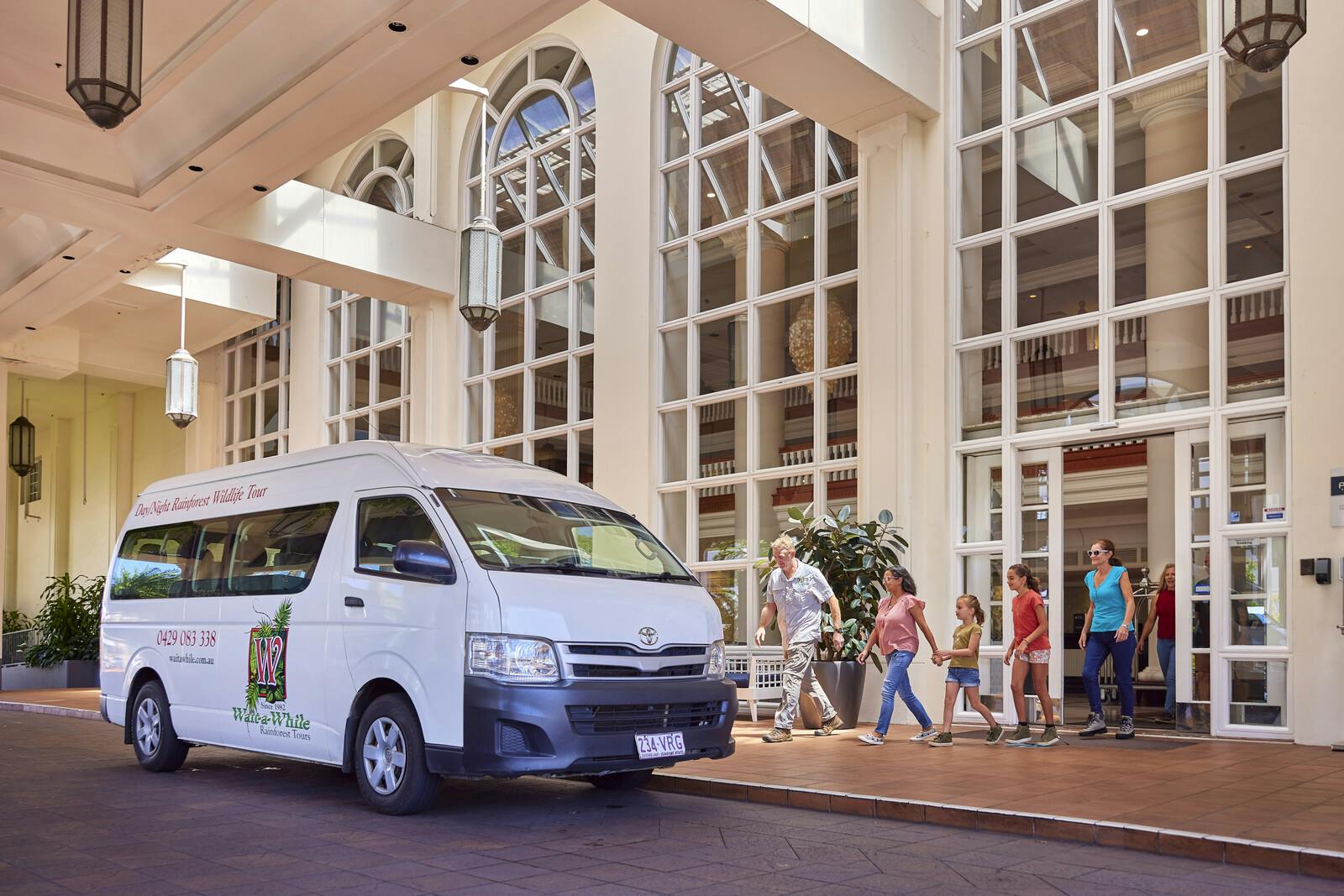 大きな窓のある建物の前に白いバンが駐車されており、大人と子供を含む人々のグループが通り過ぎています。