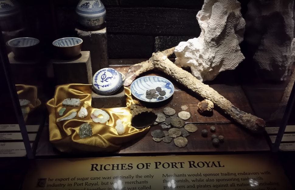 Kolekcja artefaktów, w tym monet, ceramiki i minerałów, eksponowana na złotym płótnie ze znakiem „Bogactwa Port Royal”.
