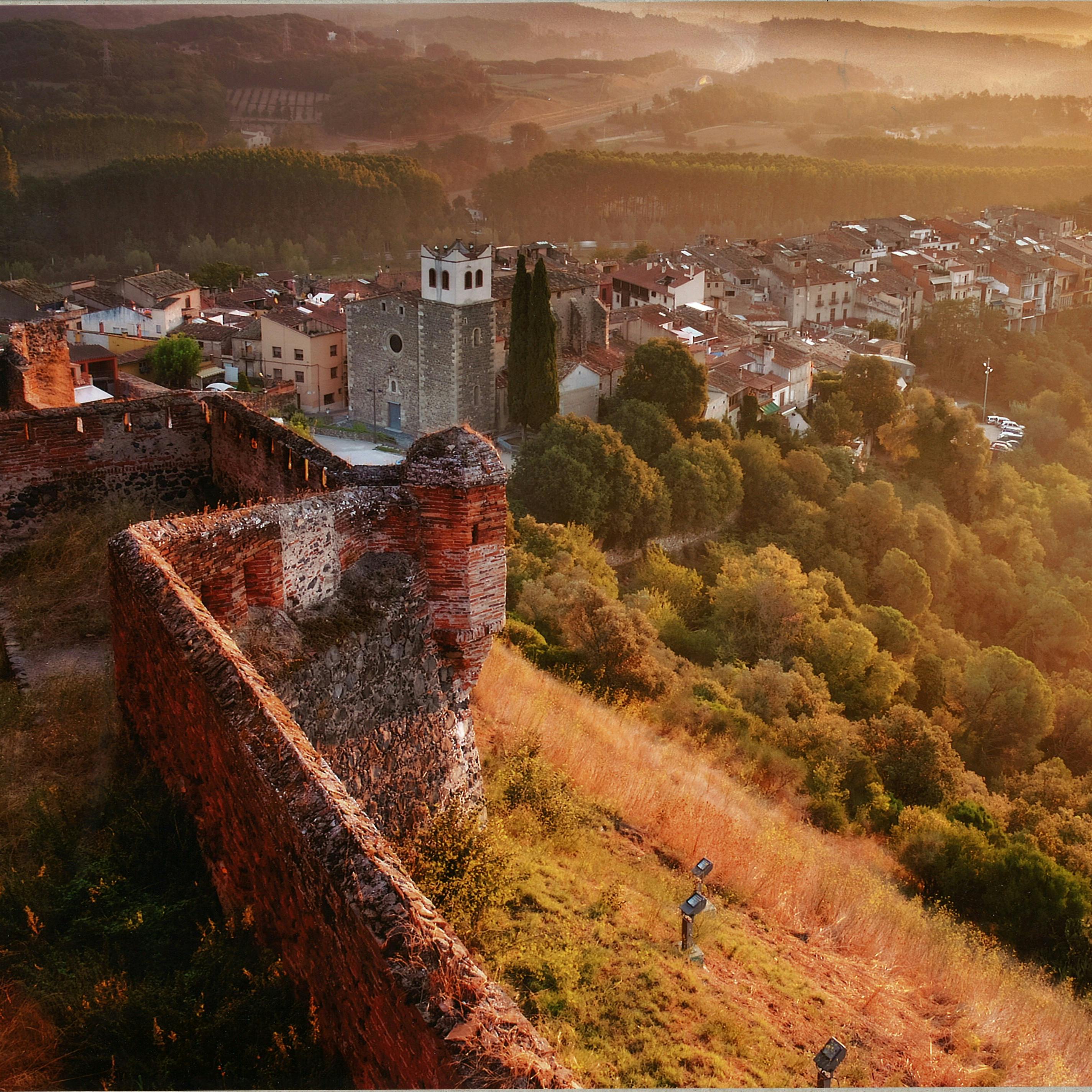A panoramic view of a rustic town at sunset, featuring an old stone fort, buildings, trees, and hilly terrain in the background.