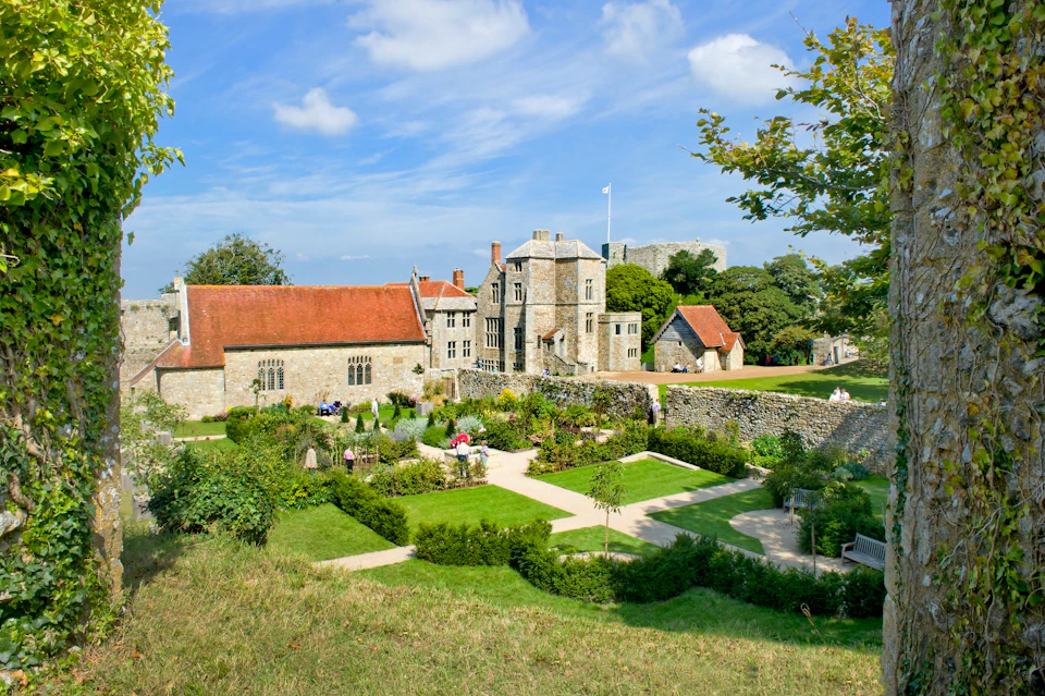 A historic stone manor with red roofs, surrounded by manicured gardens and pathways under a blue sky with scattered clouds.