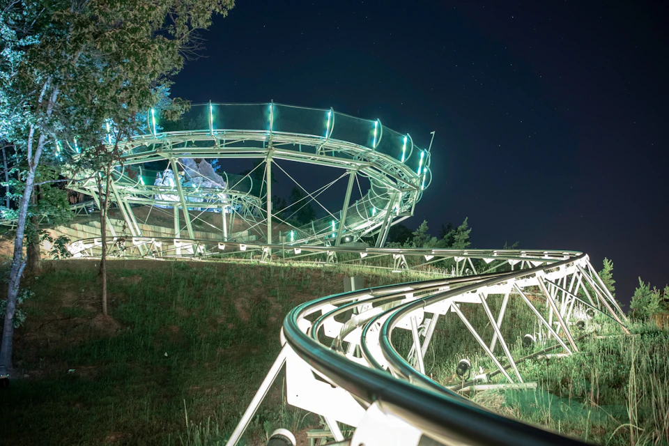 A rollercoaster track winding through a grassy area at night, leading up to an illuminated circular section. Trees in the background.