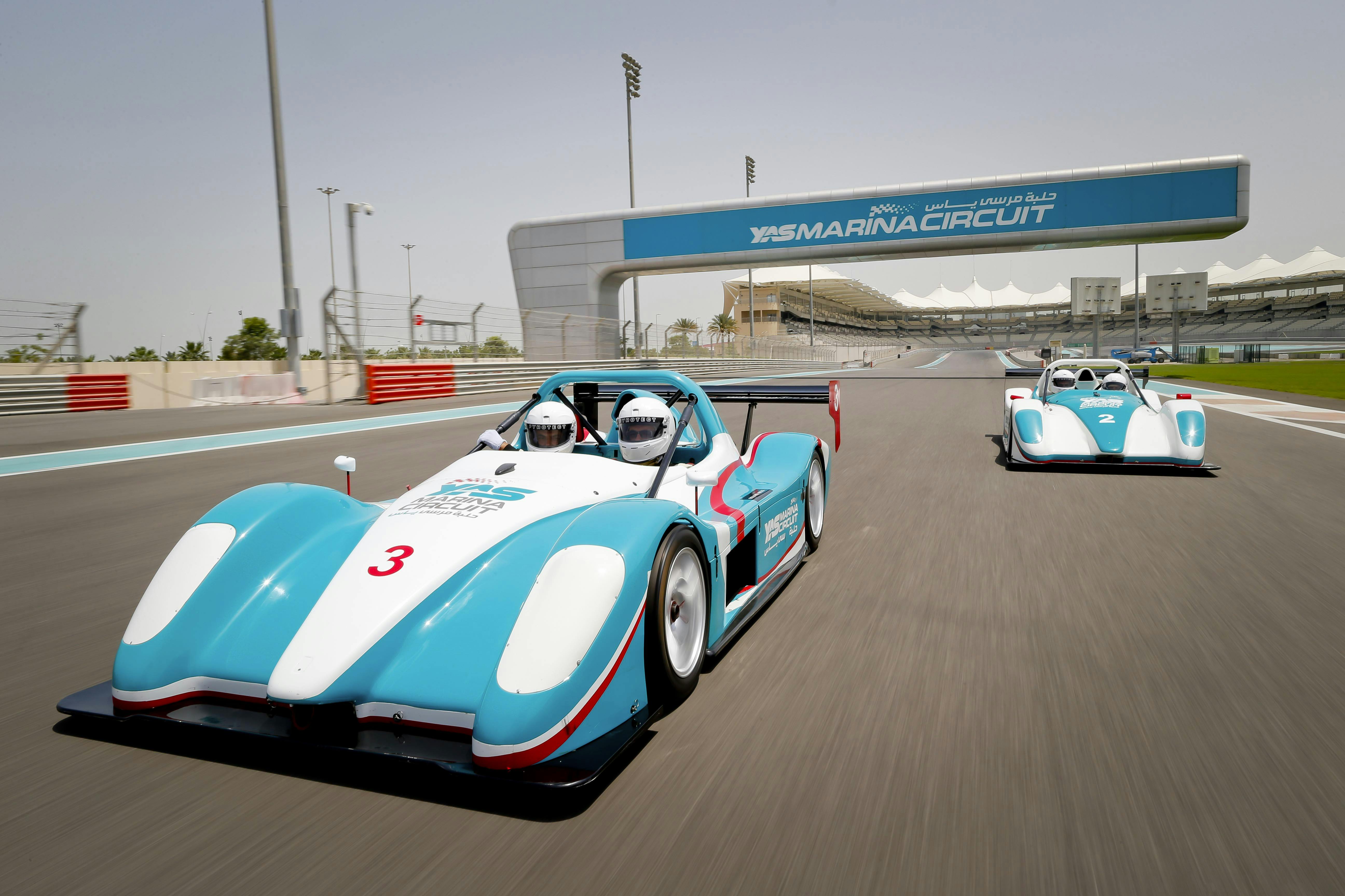 Two race cars driving on Yas Marina Circuit track under an overpass, with clear skies and a distant grandstand in the background.