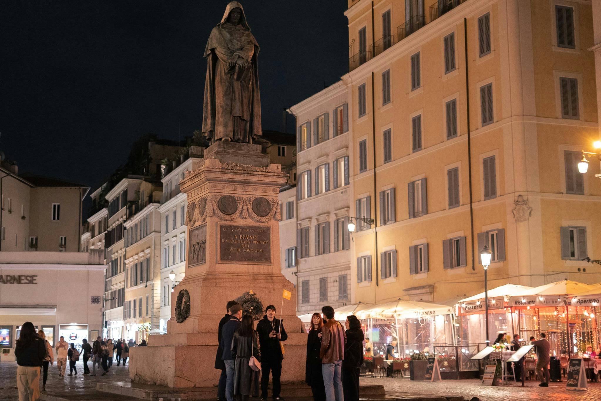 A group of people gathered near a statue in a European plaza at night, surrounded by lit building facades and storefronts.