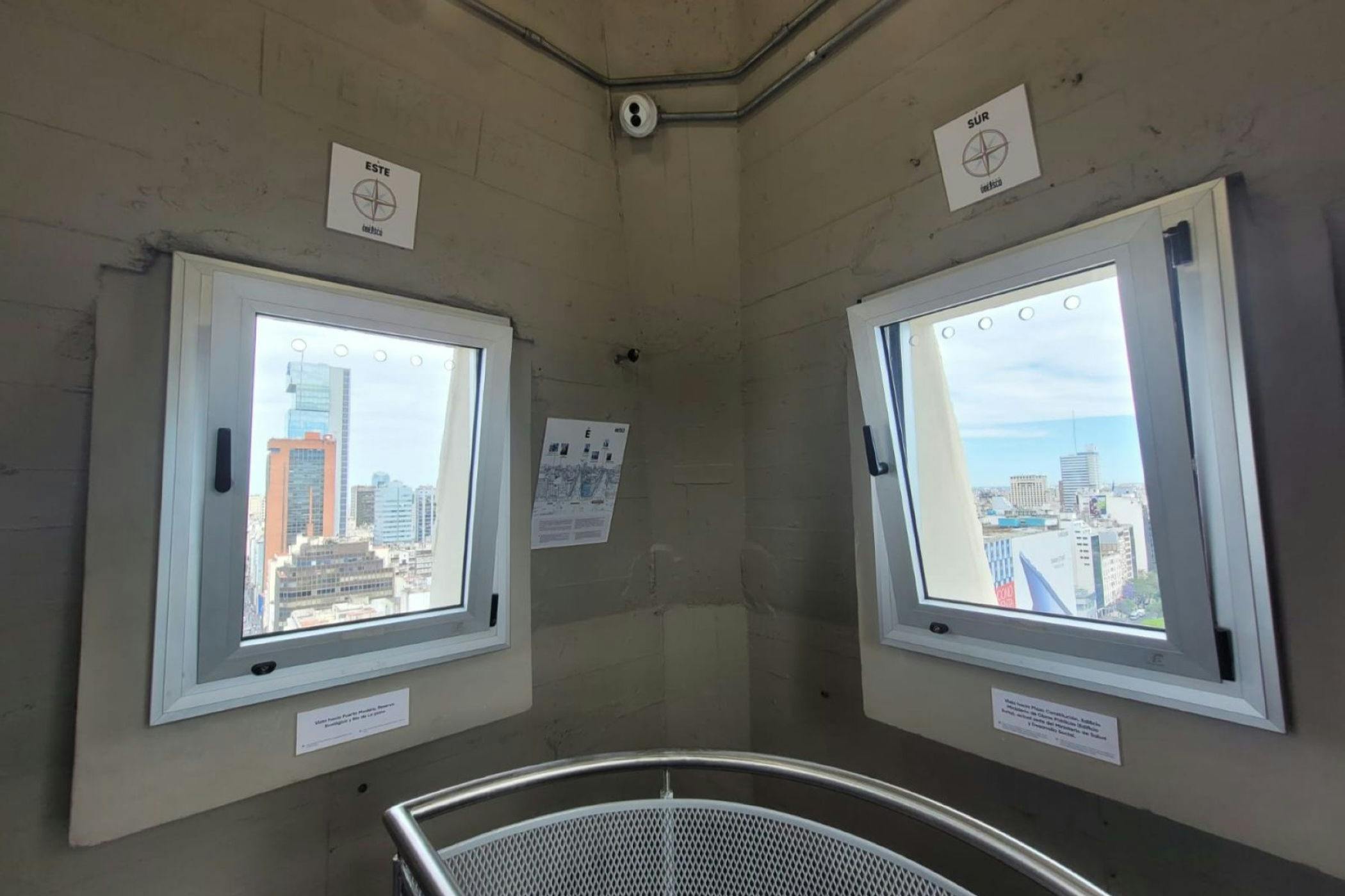 Interior view of a tower corner with two windows showing a cityscape, directional signs, and a railing in the foreground.
