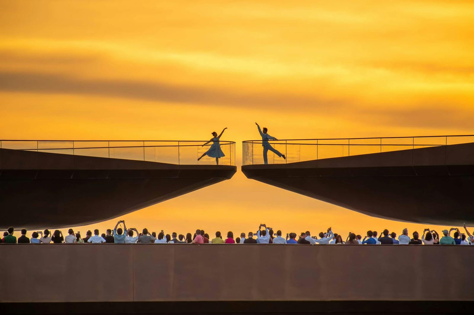 Two dancers in silhouette perform on a bridge at sunset, observed by a crowd below.