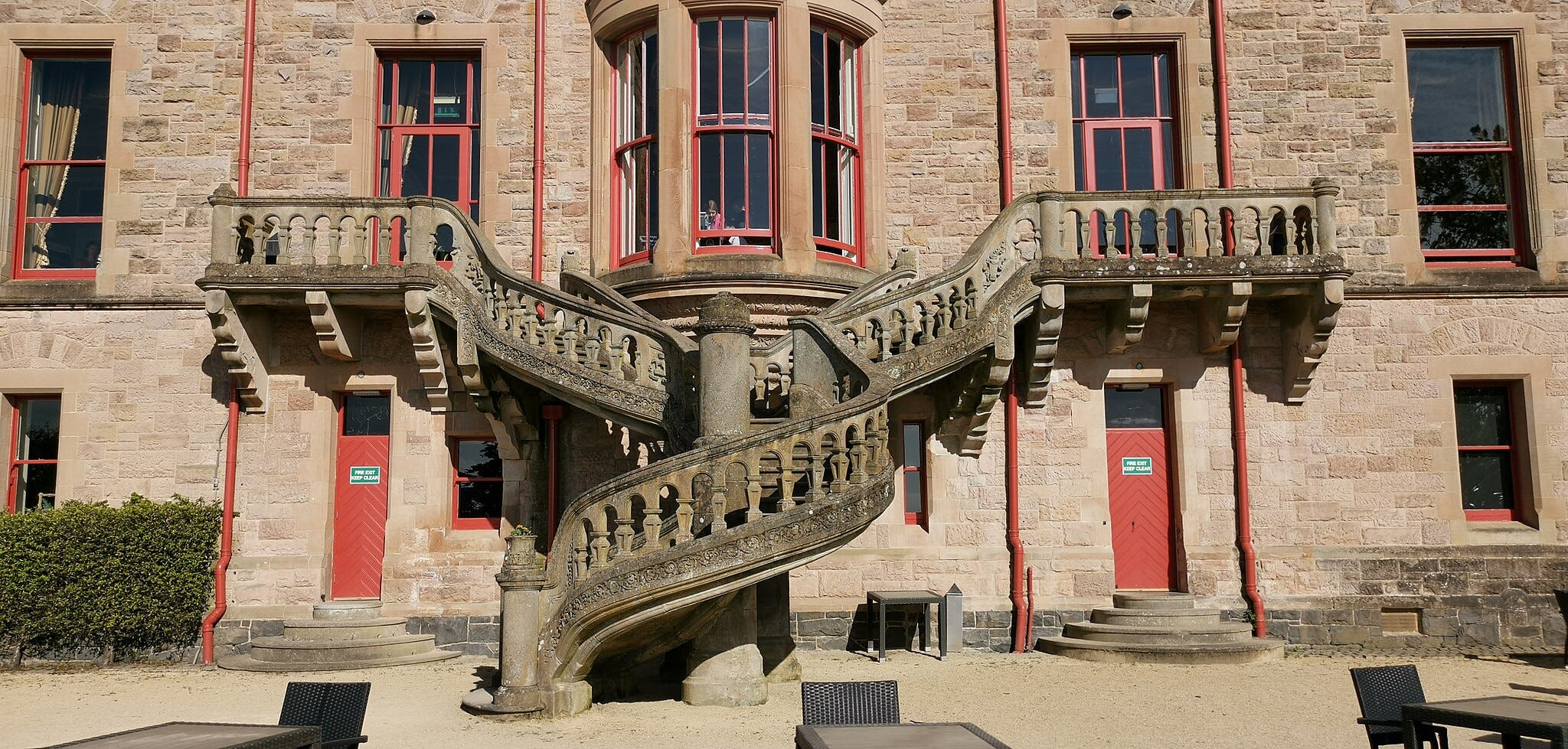 Stone building with twin decorative staircases, red-framed windows, and a red door labeled "FIRE EXIT KEEP CLEAR."