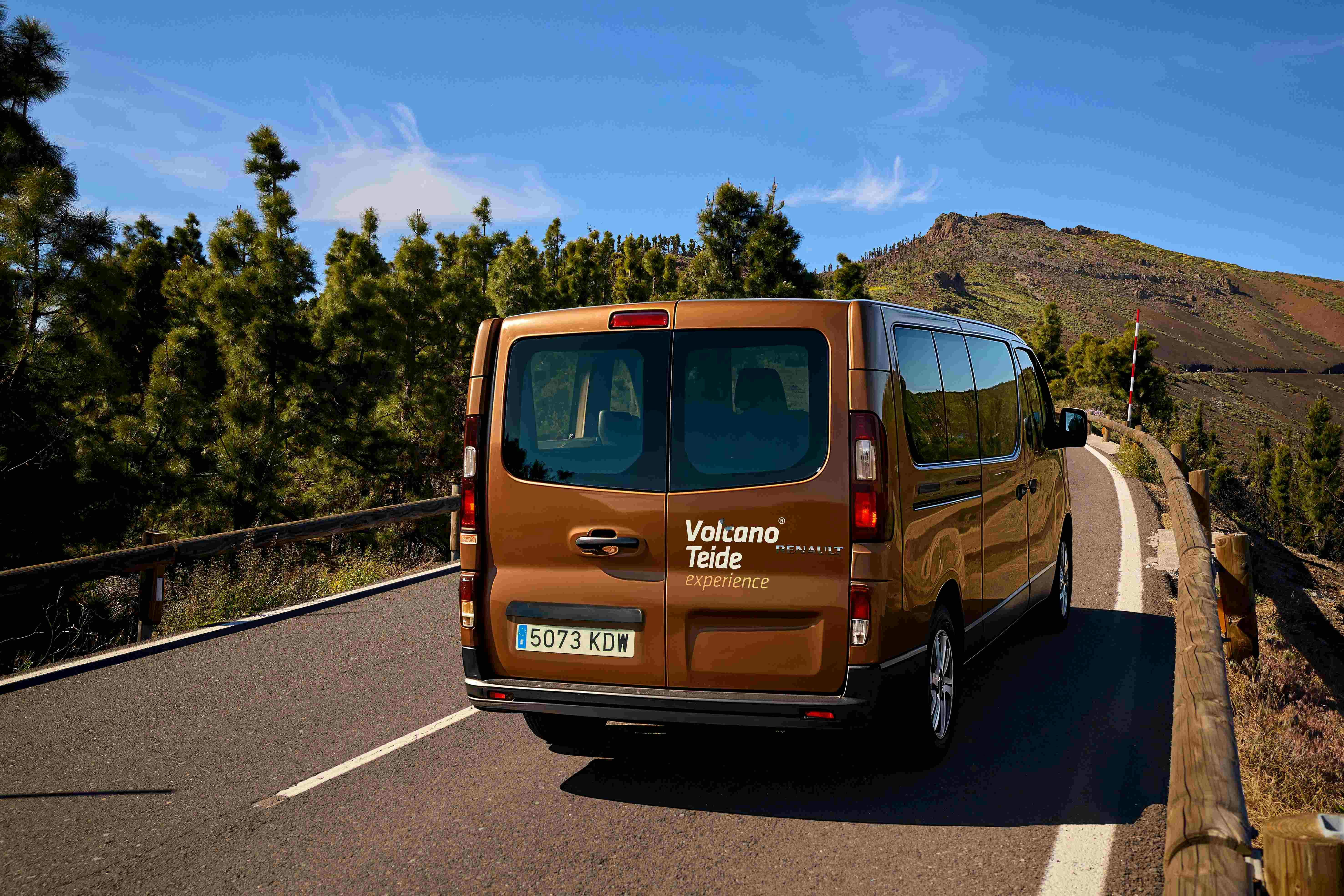 A brown van with "Volcano Teide experience" branding drives on a mountain road surrounded by trees and hills under a clear sky.