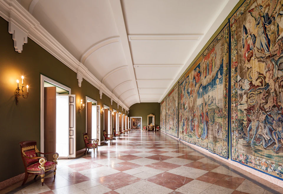 Elegant hallway with high arched ceiling, checkered floor, ornate wall tapestry, and vintage chairs along the walls.