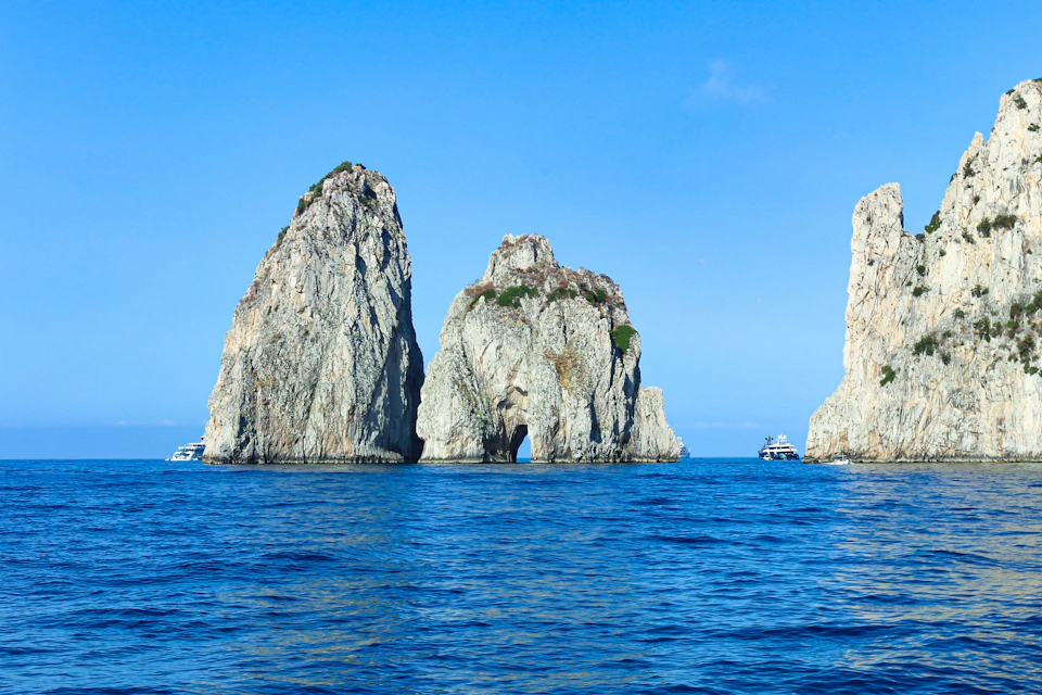 Three large rocky sea stacks with vegetation, surrounded by blue water, under a clear blue sky. Boats are visible nearby.