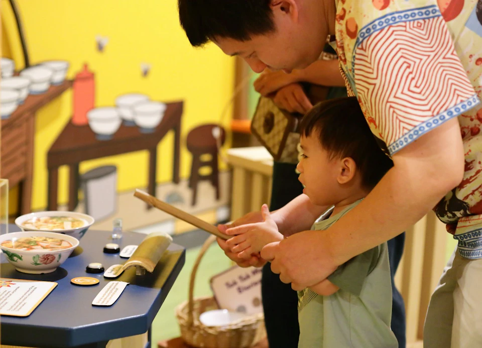 A child holding a drumstick assisted by an adult in an interactive exhibit with a vibrant indoor setting.