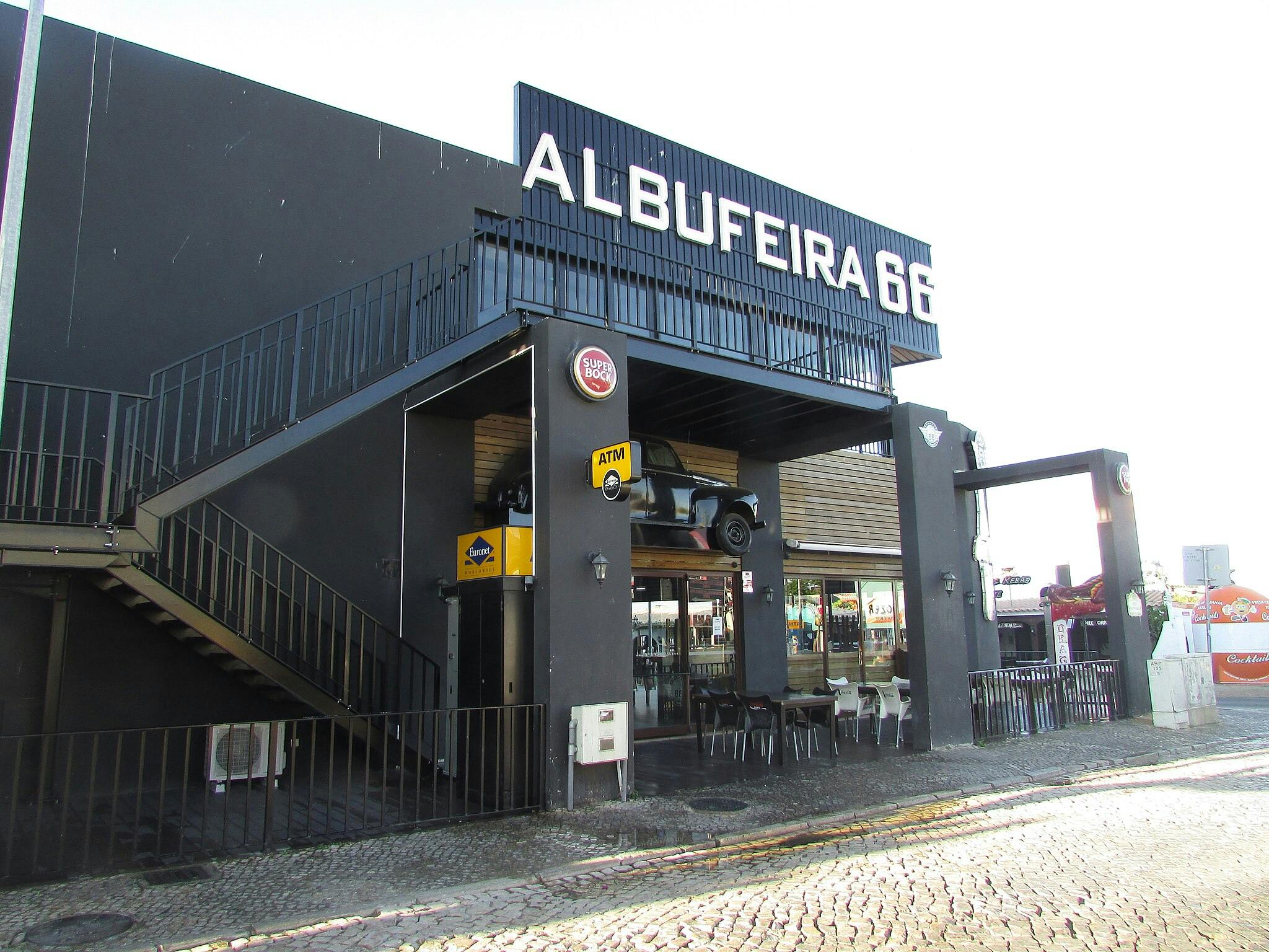 Exterior of a building with "ALBUFEIRA 66" signage, an ATM, stairs, and a car mounted above an outdoor seating area on a cobblestone street.