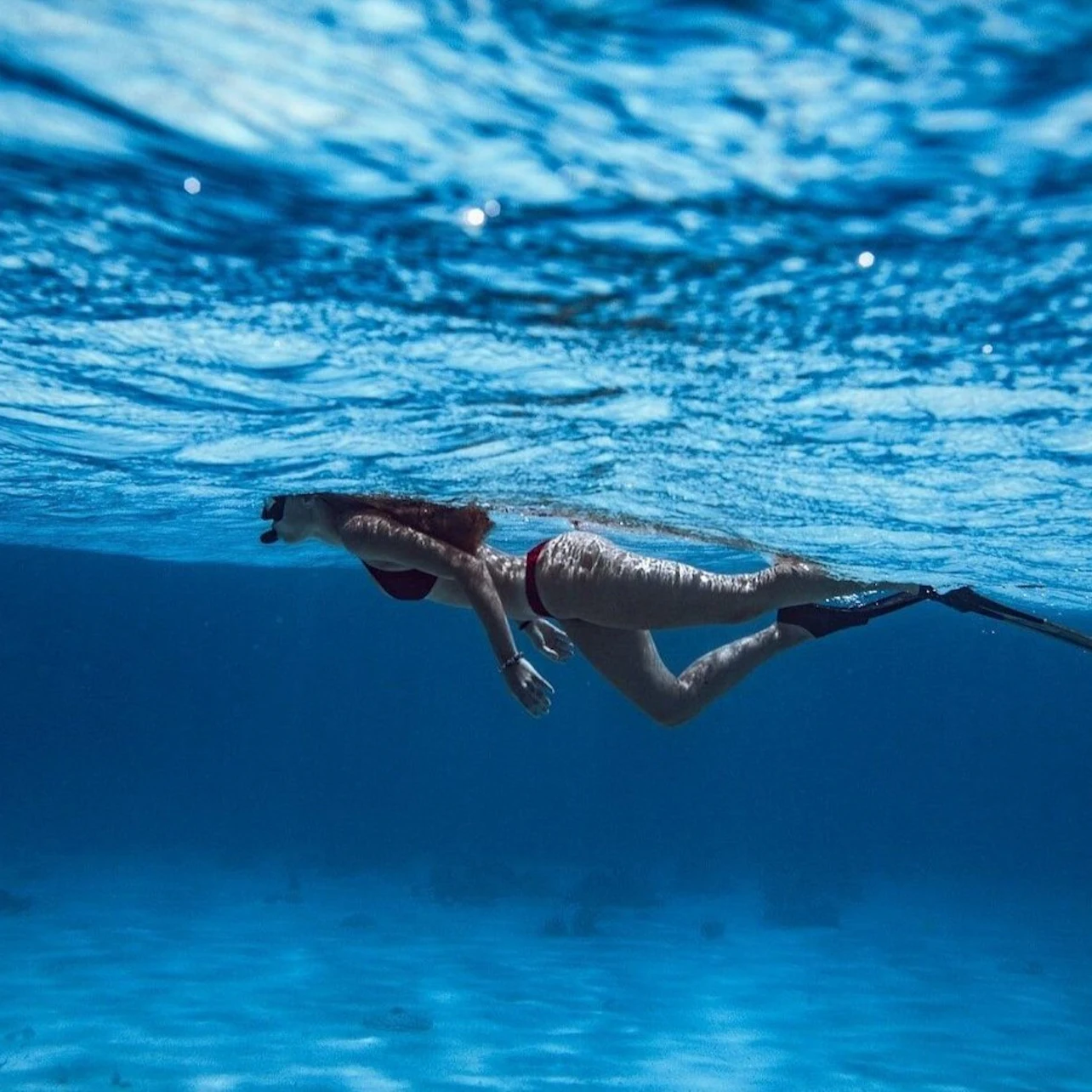 A person swimming underwater in a clear blue sea, wearing fins and a swimsuit, with sunlight filtering through the water.