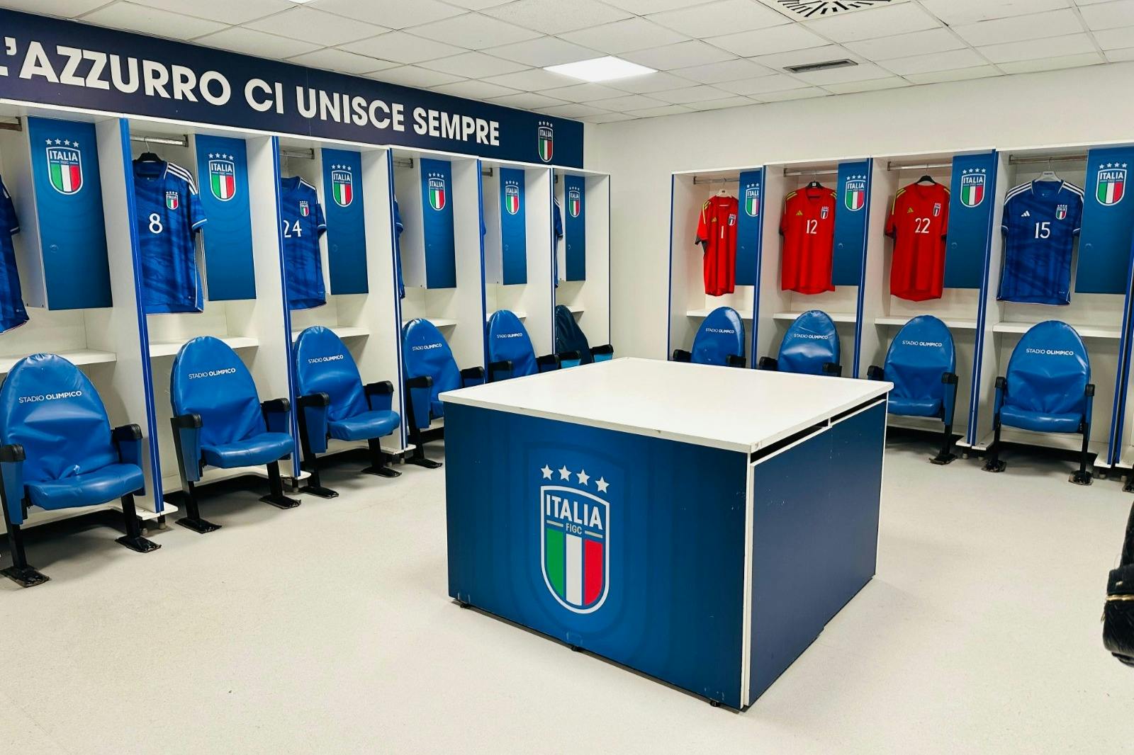 Spogliatoio con maglie da calcio italiane rosse e blu in esposizione, sedili blu e un tavolo centrale con il logo della squadra di calcio italiana.