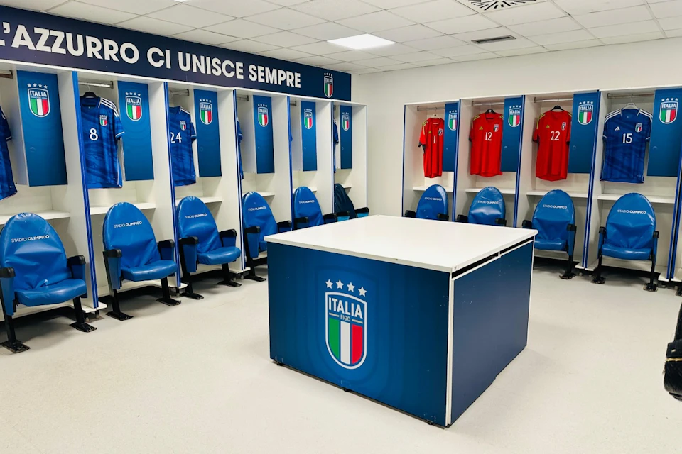 Locker room with red and blue Italian soccer jerseys on display, blue seats, and a central table featuring the Italian soccer team logo.