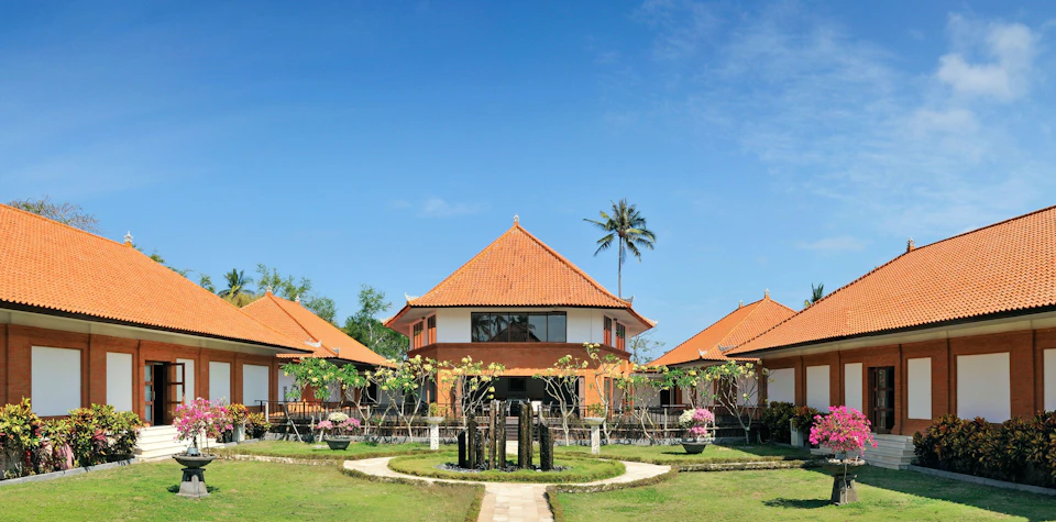A large, orange-roofed building with surrounding smaller structures, lush greenery, and a clear blue sky in the background.