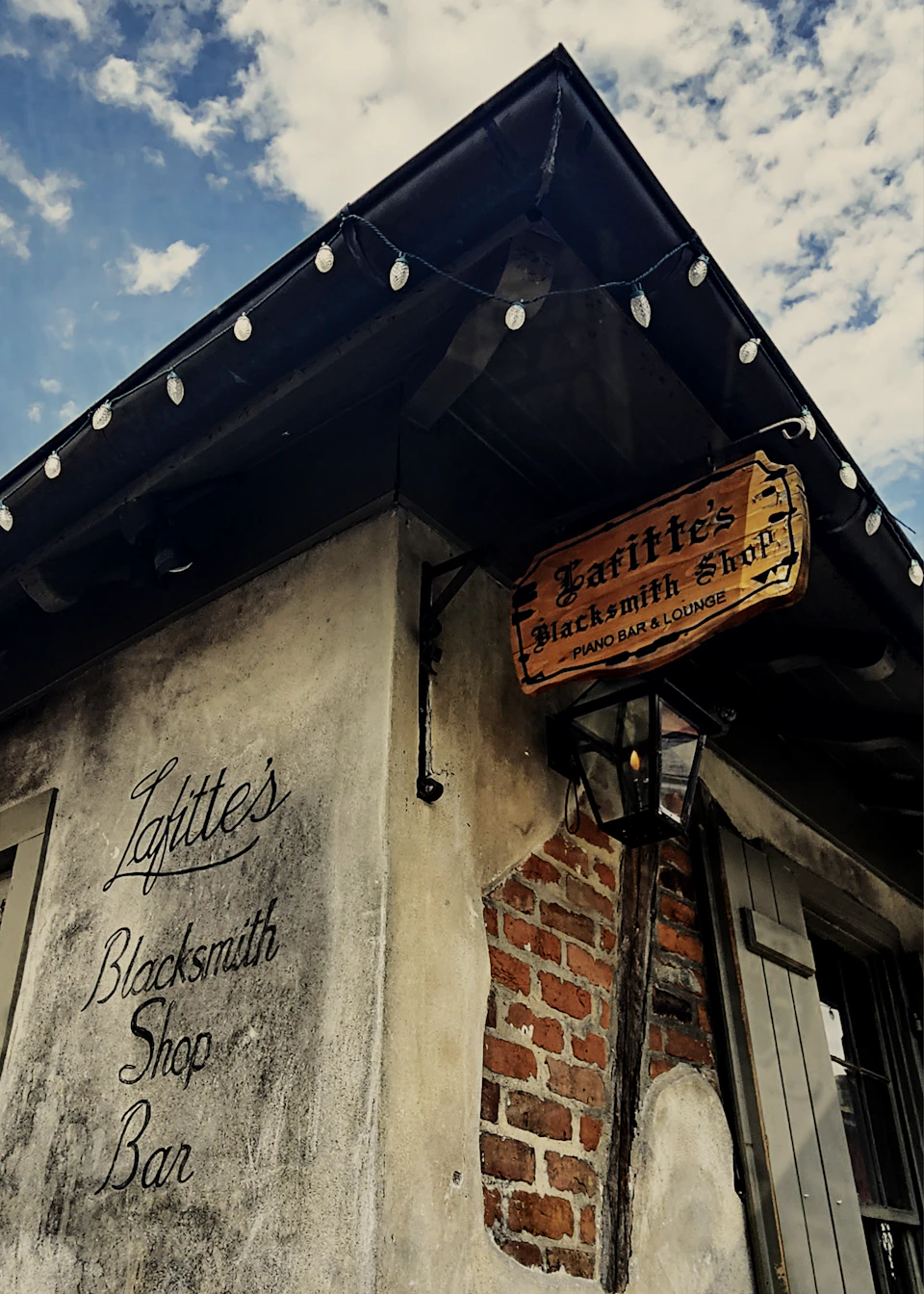 Exterior of Lafitte's Blacksmith Shop with a hanging sign and string lights under a partly cloudy sky.