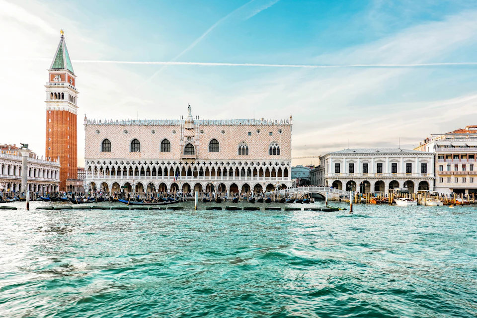 Ornate historical building with arched windows and columns on a waterfront, boats docked in front, under a clear blue sky.