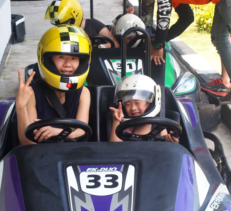 Two people in helmets sitting in go-kart number 33, both making peace signs. Others are visible in the background.