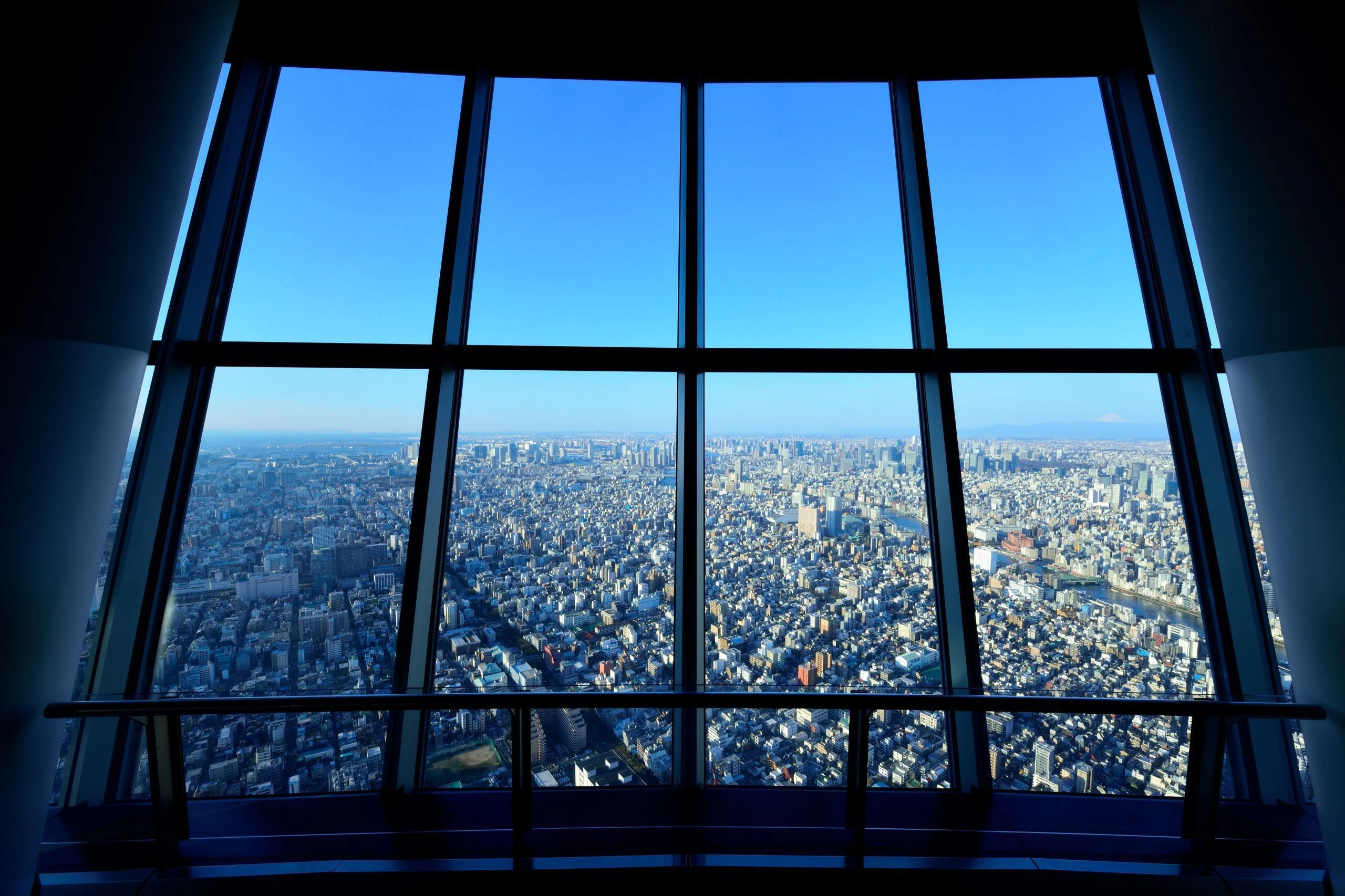 Aerial view of a densely packed cityscape seen through large windows on a high floor, with a clear blue sky.