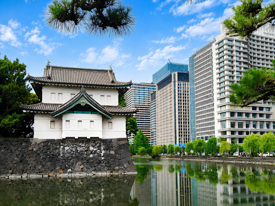 A traditional Japanese building by a moat, with modern skyscrapers in the background under a blue sky.