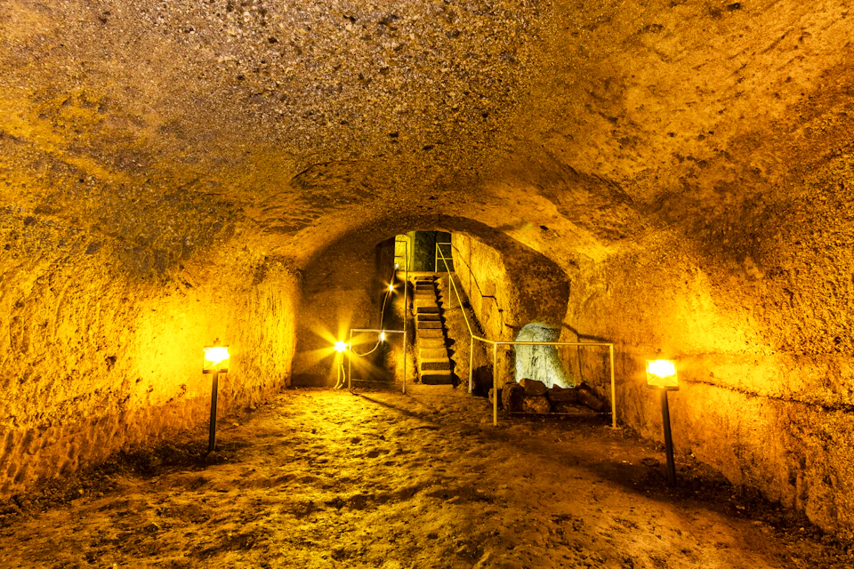 Underground cave with lit lanterns, rough walls, and a staircase leading to an upper level. The area is illuminated with a warm glow.