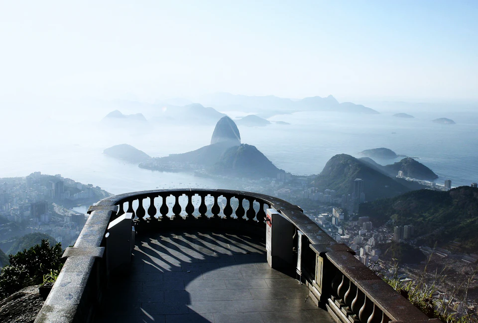 Stone balcony overlooking a cityscape with mountains and water, featuring a prominent rock formation in the distance.