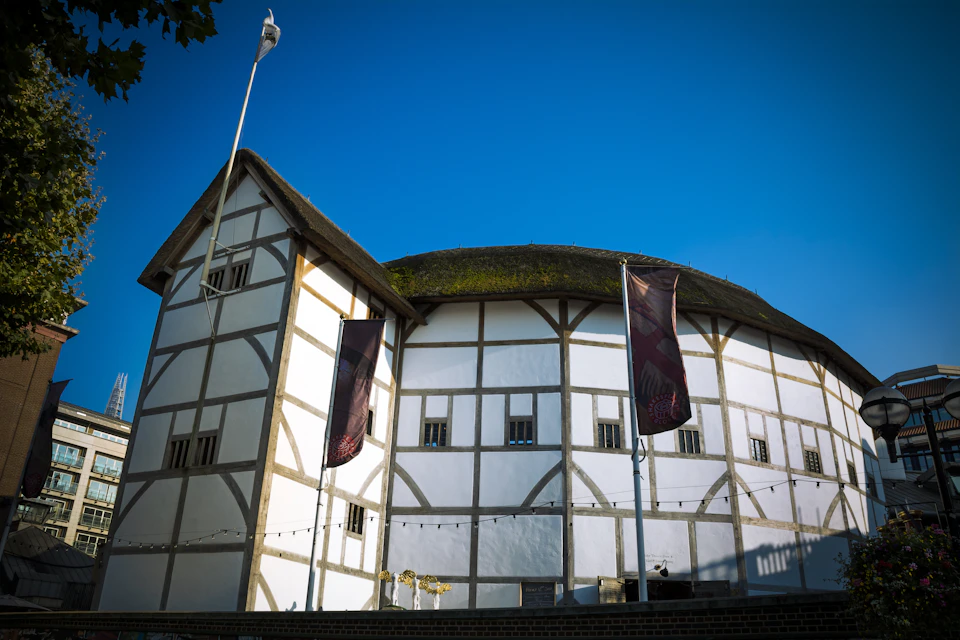 Round white building with timber frames and a thatched roof, flanked by two tall flags beside it, against a clear blue sky.