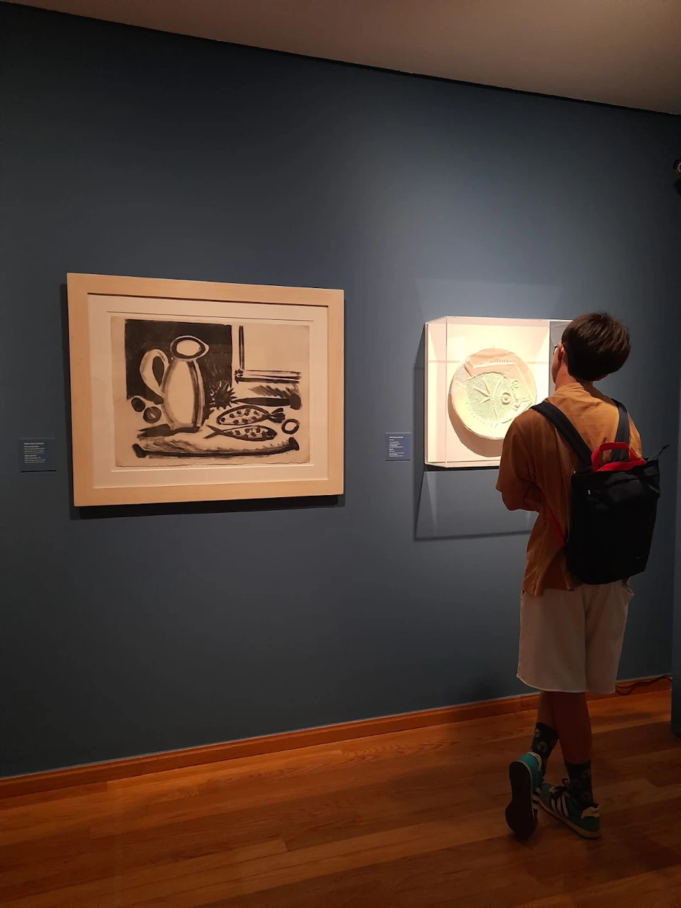 Person with a backpack observing two framed artworks on a blue museum wall; one is a black and white drawing, the other a circular piece.
