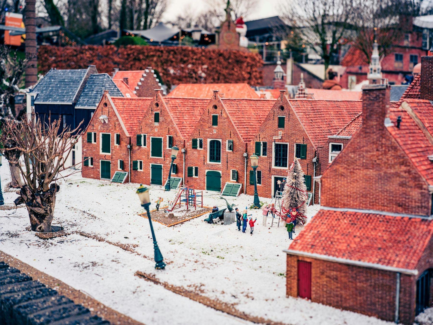 雪に覆われた地面、レンガ造りの家、遊び場、人形が置かれたモデルビレッジ。 背景には木と生垣が表示されます。