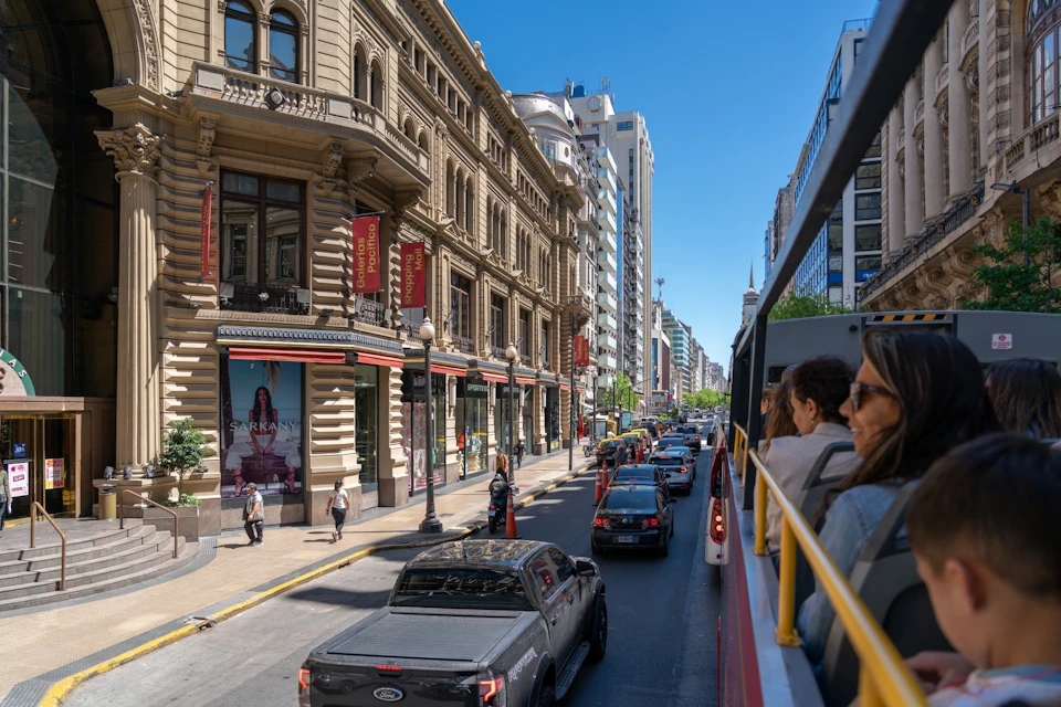 A street in a city with traffic, pedestrians, and historic buildings, viewed from a double-decker bus on a sunny day.