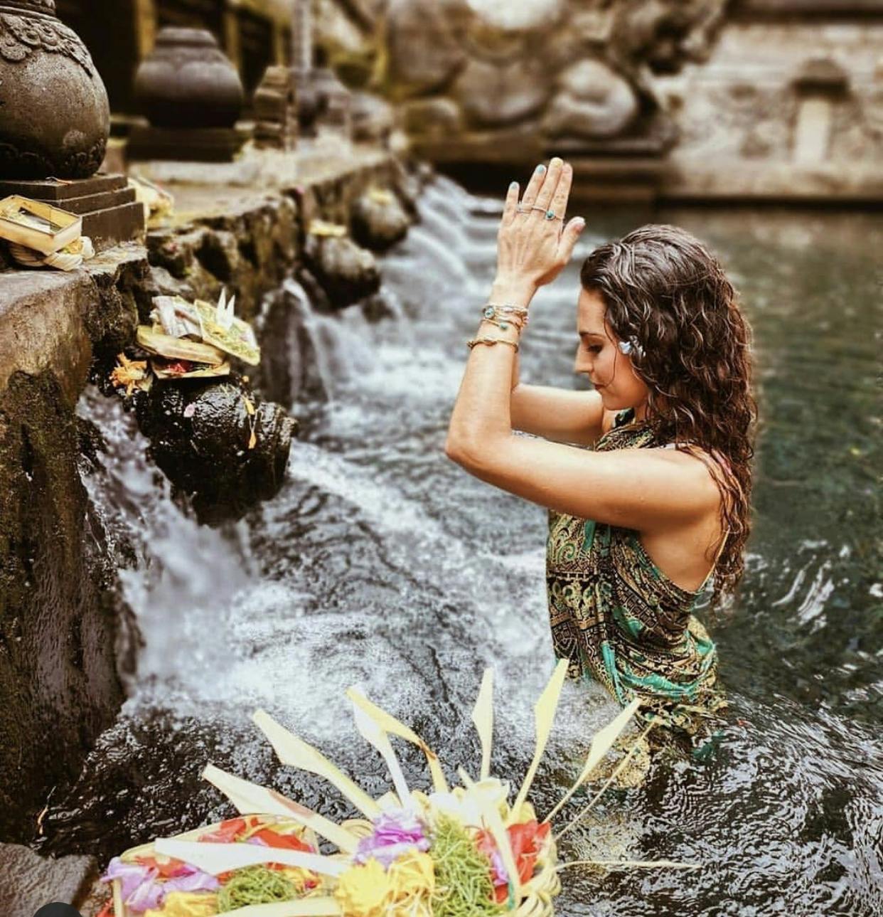 A woman stands in a stone pool, hands in prayer, near flowing water and offerings.