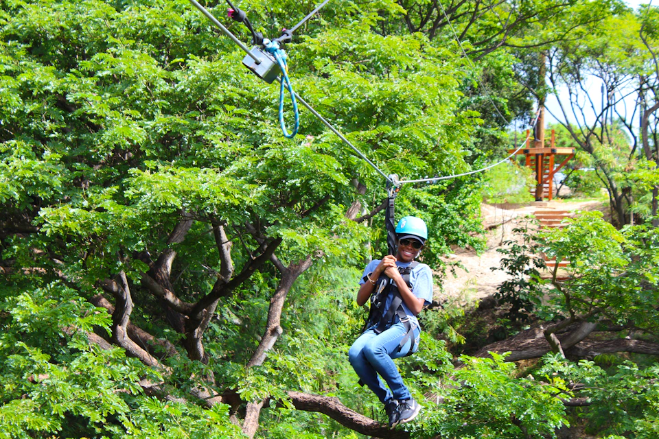 A person wearing a helmet and harness rides a zipline through a lush, green treetop canopy.