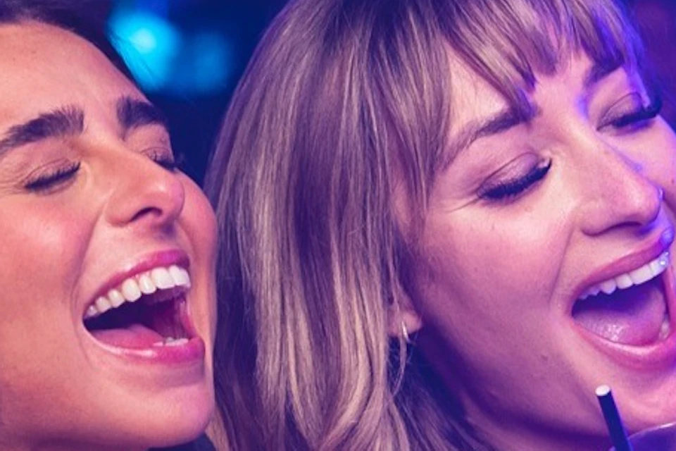 Two people smiling and singing in a lively nightclub setting, holding drinks, surrounded by colorful lights and other patrons.