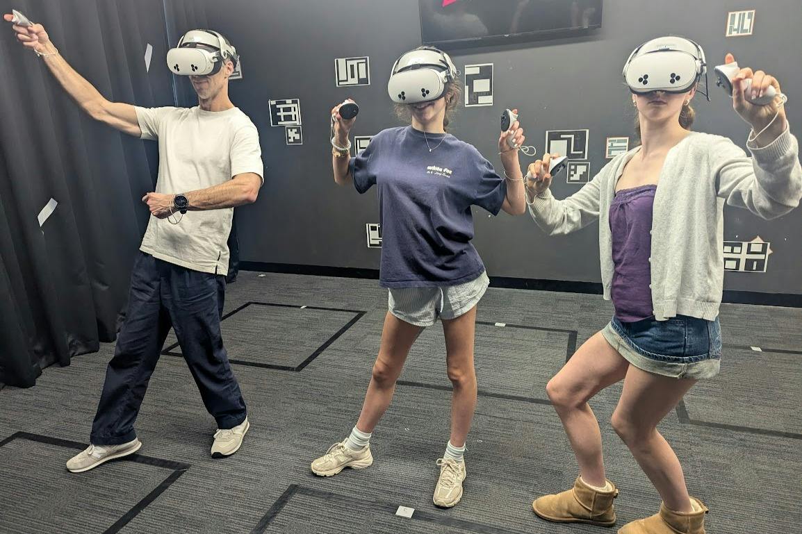 Duas pessoas com óculos de realidade virtual e controladores nas mãos, de pé numa sala com paredes cinzentas e decorações estampadas.