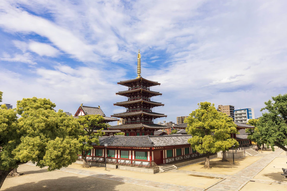 A traditional Japanese pagoda surrounded by trees under a partly cloudy sky, with modern buildings in the background.