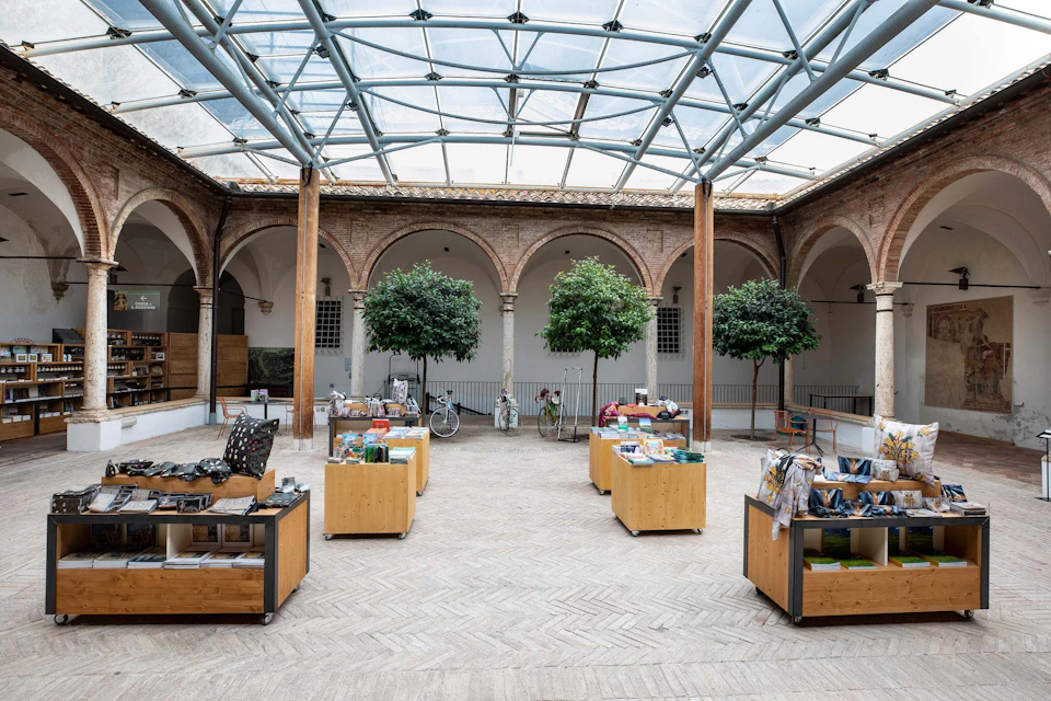 An open courtyard with a glass roof, surrounded by arches and columns, contains several wooden display tables and potted trees.