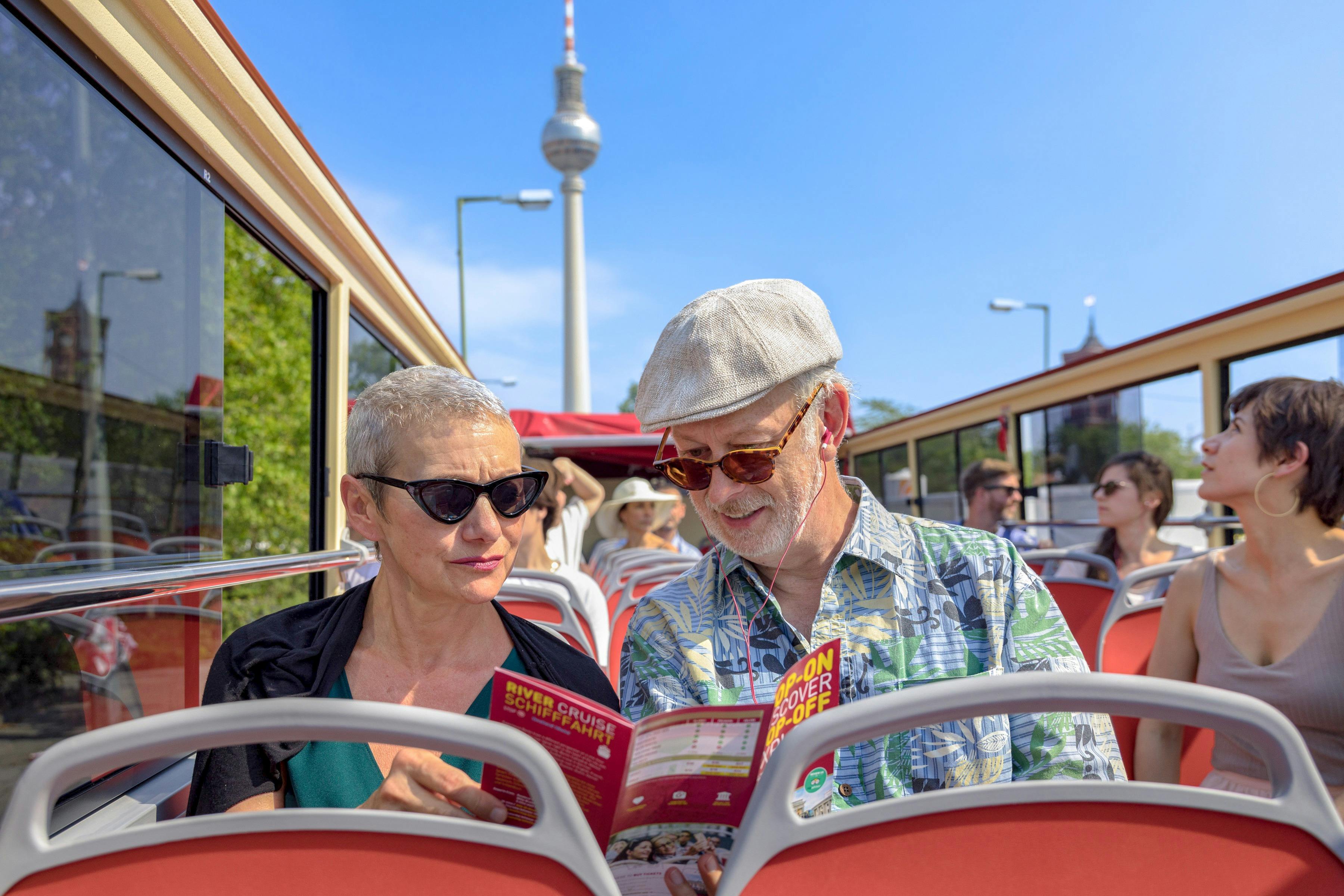 Zwei Personen, die auf dem Oberdeck eines Sightseeing-Busses sitzen und Broschüren lesen. Im Hintergrund ist der Berliner Fernsehturm zu sehen.